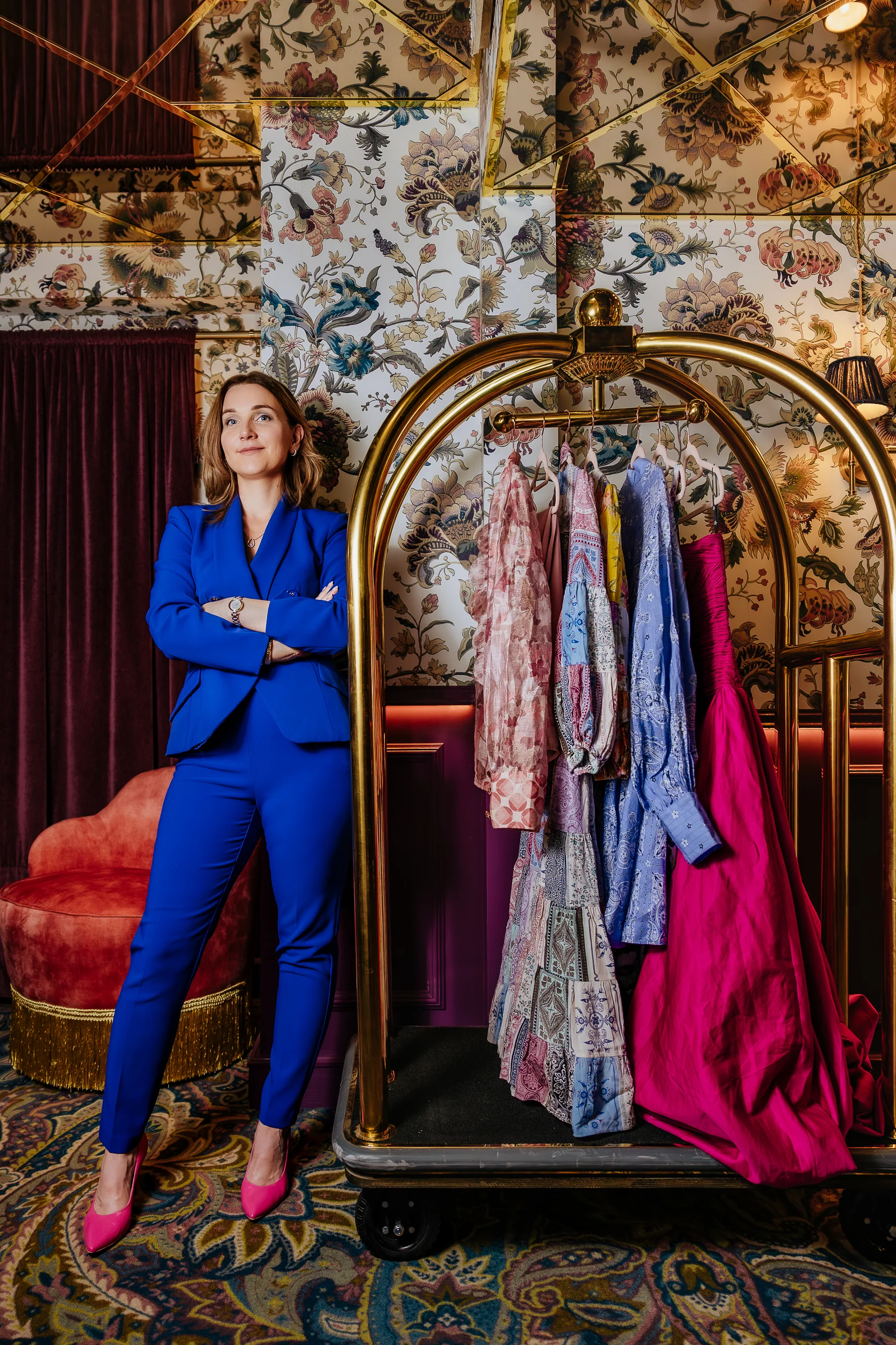 Ekspertka kolorów Dorota Janisz w kobaltowym garniturze i eleganckim wnętrzu stoi przy wieszaku ubrań. 